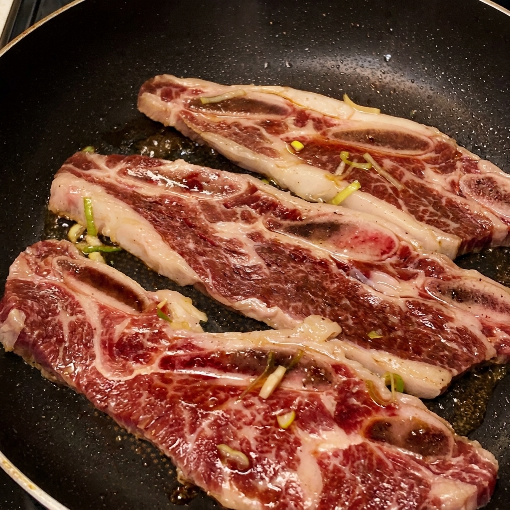 Trois travers de bœuf grésillant dans une poêle avec la marinade sombre qui bouillonne et se réduit en un glaçage épais et brillant.