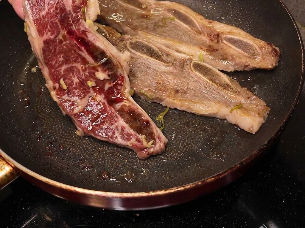 Une main retournant un travers de bœuf partiellement cuit dans une poêle, révélant une surface caramélisée et bien dorée.