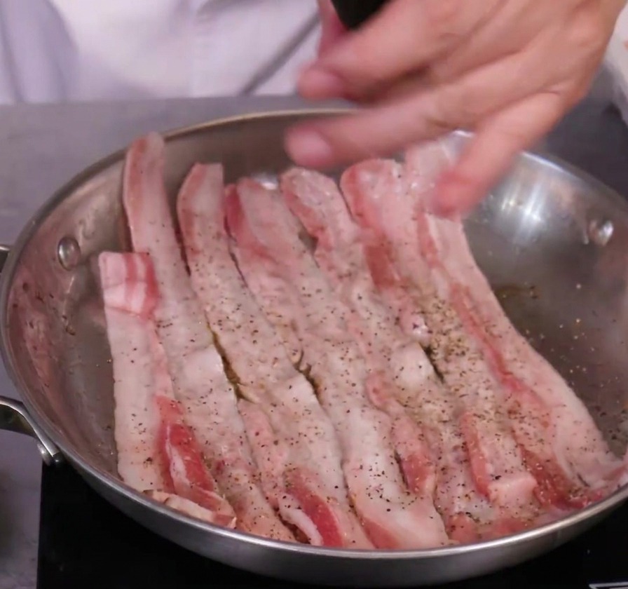 Assaisonnement de tranches de poitrine de porc avec un moulin à poivre noir dans une poêle en acier.