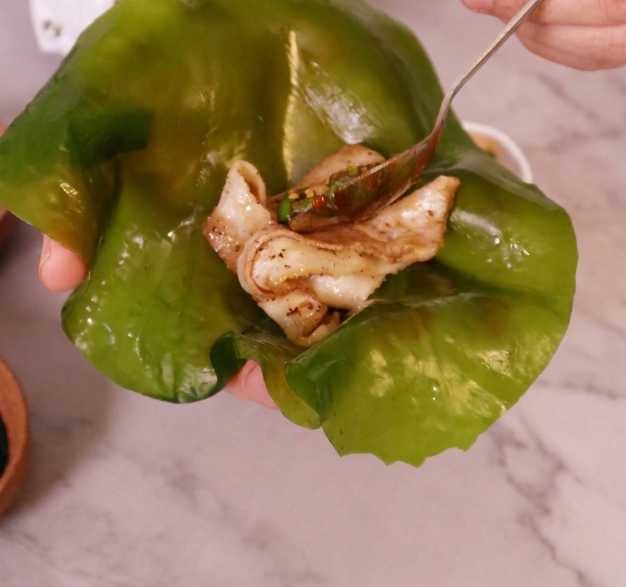 Une main tenant une grande feuille de varech vert translucide avec un morceau de poitrine de porc cuite posé au centre.
