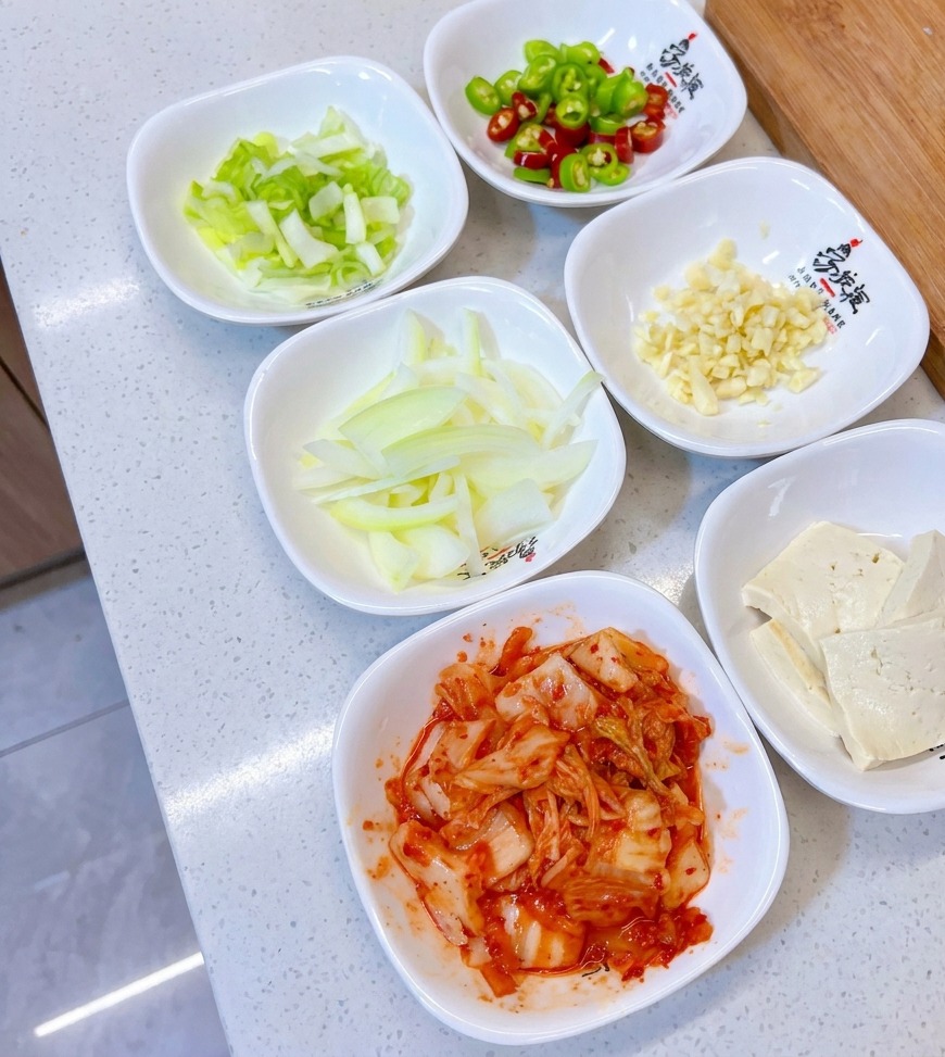 Un assortiment de petits bols blancs contenant des oignons verts émincés, des piments verts et rouges, de lail haché, des oignons émincés, du tofu et du kimchi.