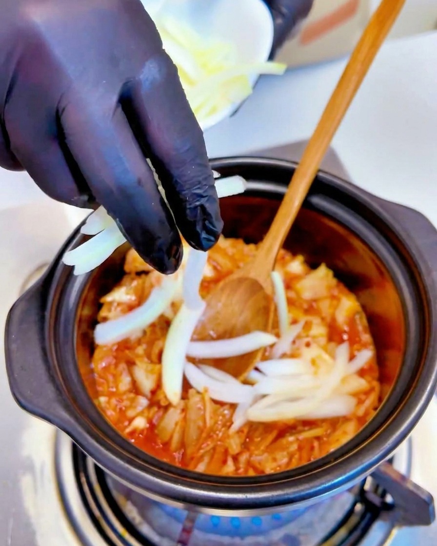 Des oignons blancs émincés ajoutés au mélange de kimchi frémissant dans une marmite par une main portant un gant noir.