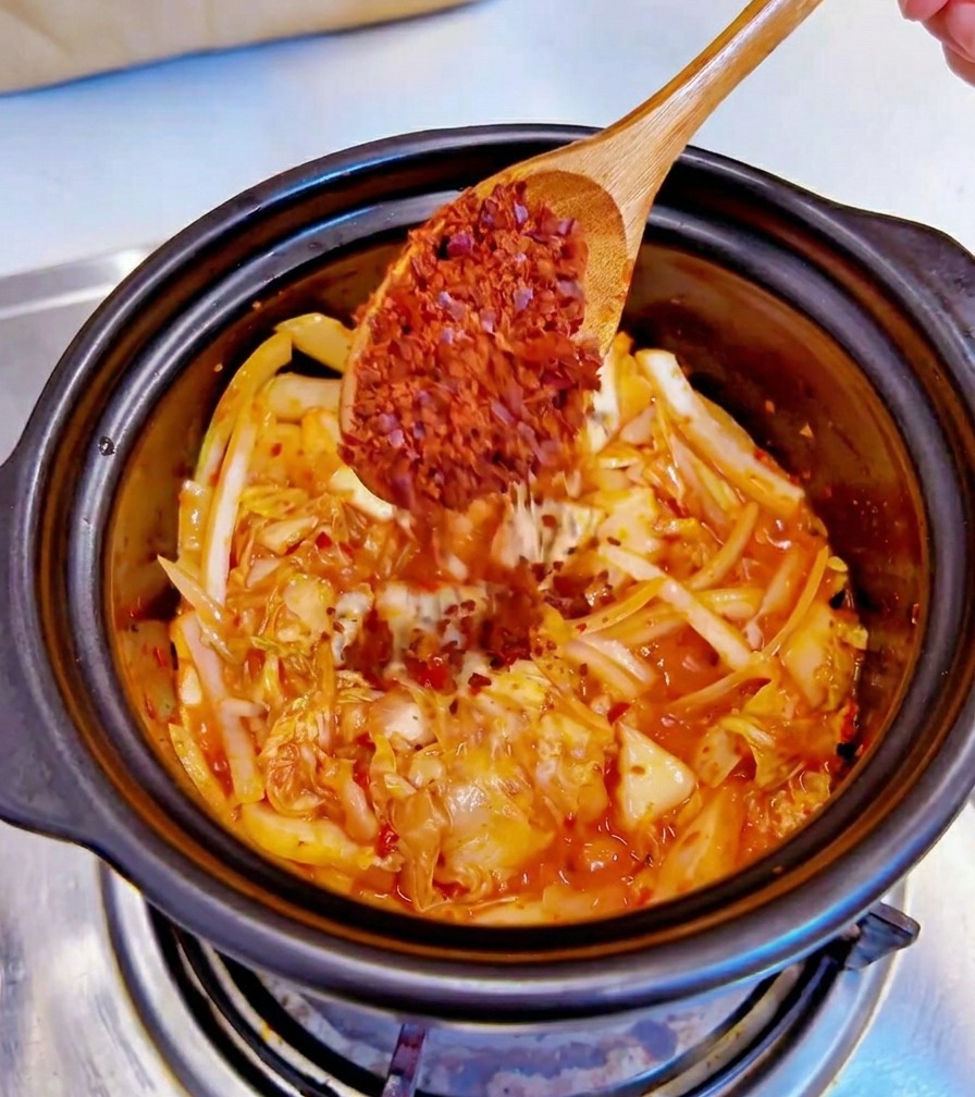 Une cuillère en bois ajoutant des flocons de piment rouge grossiers dans une marmite noire contenant du kimchi sauté et des oignons émincés.