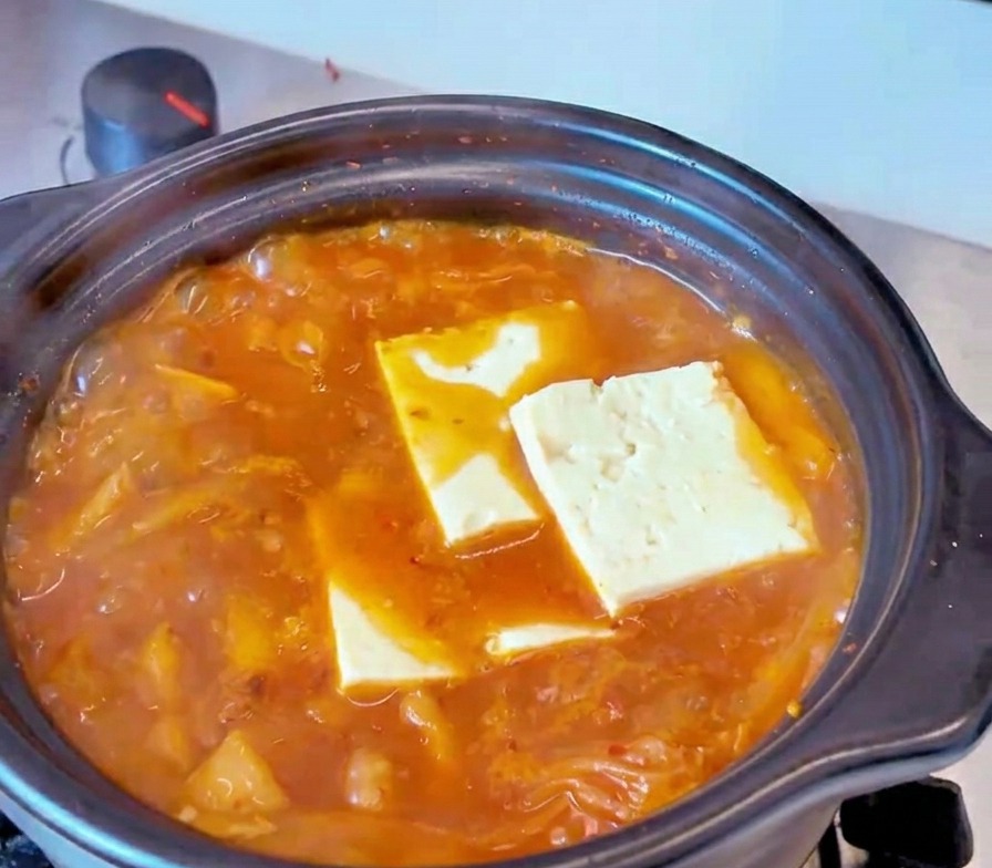 Des tranches rectangulaires de tofu blanc reposant dans un bouillon de ragoût de kimchi rouge et frémissant.