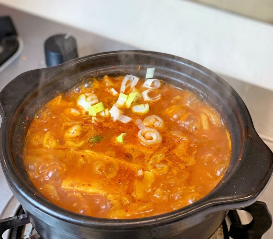 Un ragoût de kimchi au thon rouge et bouillonnant dans une marmite en terre noire traditionnelle sur une cuisinière, garni doignons verts frais et de tranches de piment.