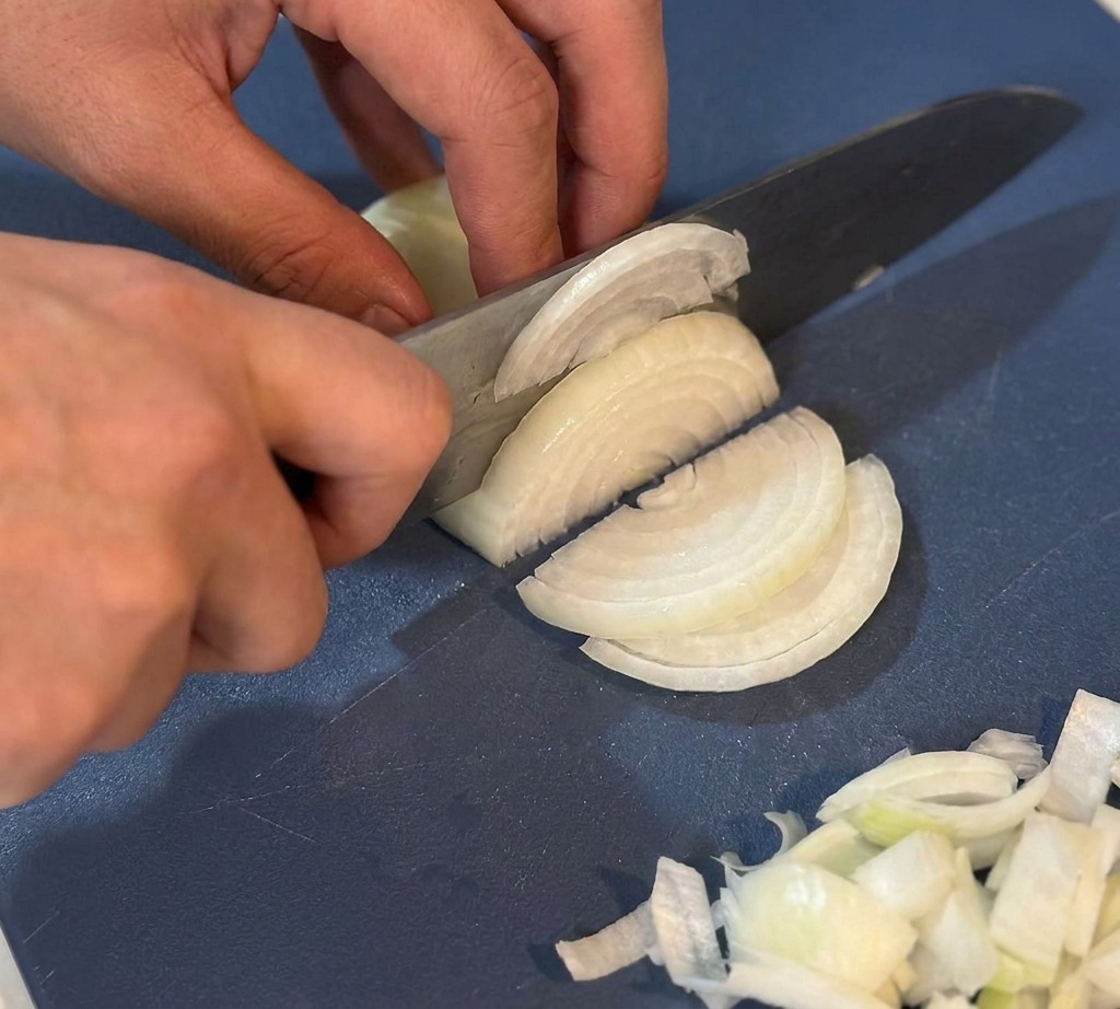 Mains utilisant un couteau de chef pour émincer finement un oignon blanc sur une planche à découper bleue.
