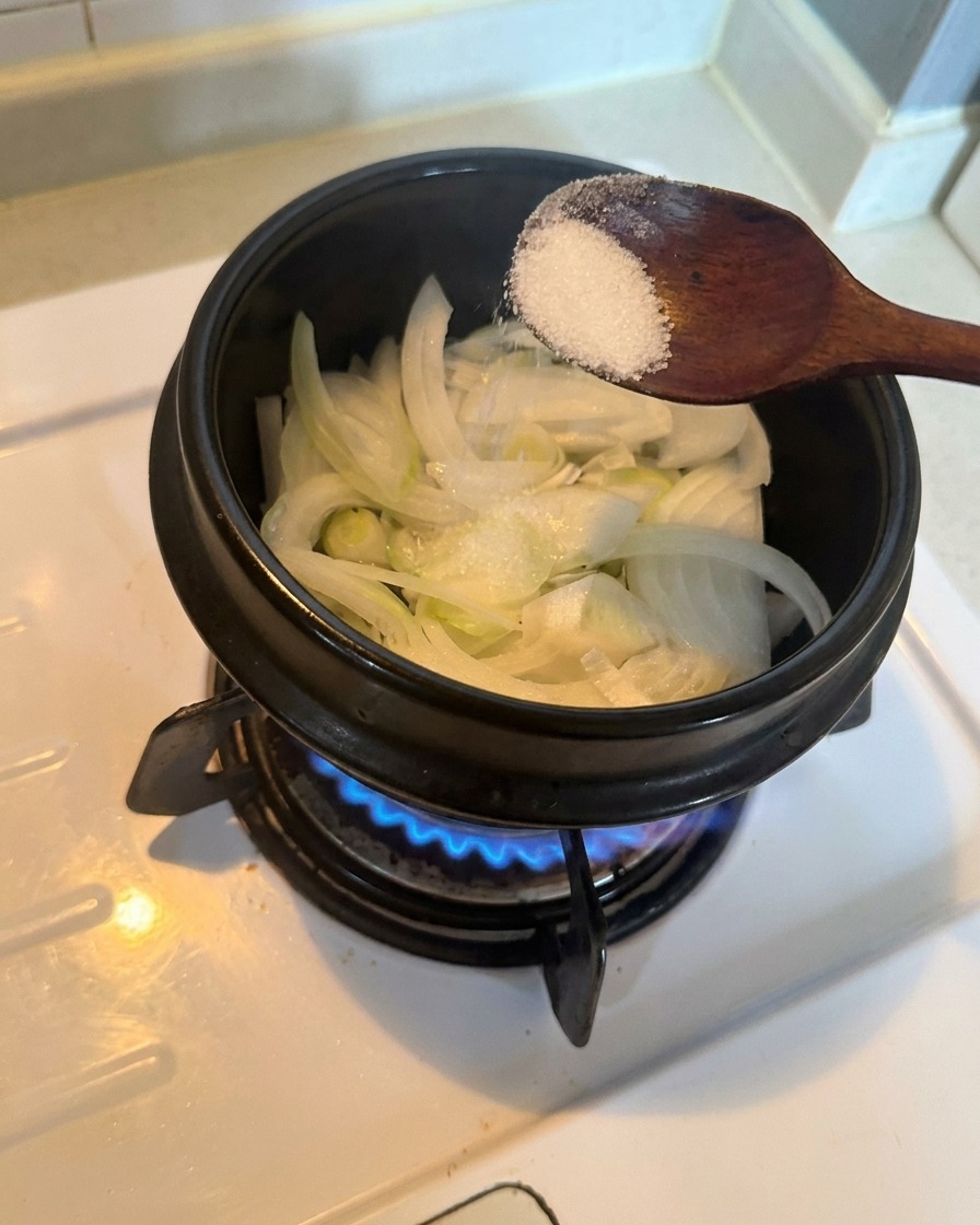 Oignons blancs émincés dans une marmite, agrémentés dune cuillère de sucre blanc au-dessus dun brûleur à gaz allumé.