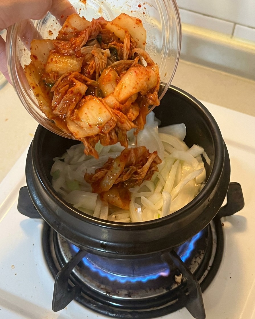 Ajout dun bol de kimchi rouge vif haché dans une marmite contenant déjà les oignons et les cébettes revenus.