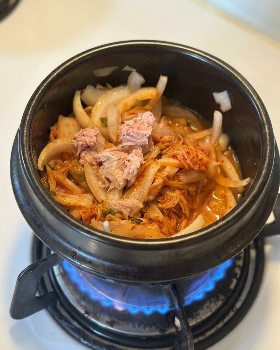 Morceaux de thon en boîte déposés sur un lit de kimchi et doignons revenus dans une marmite noire.