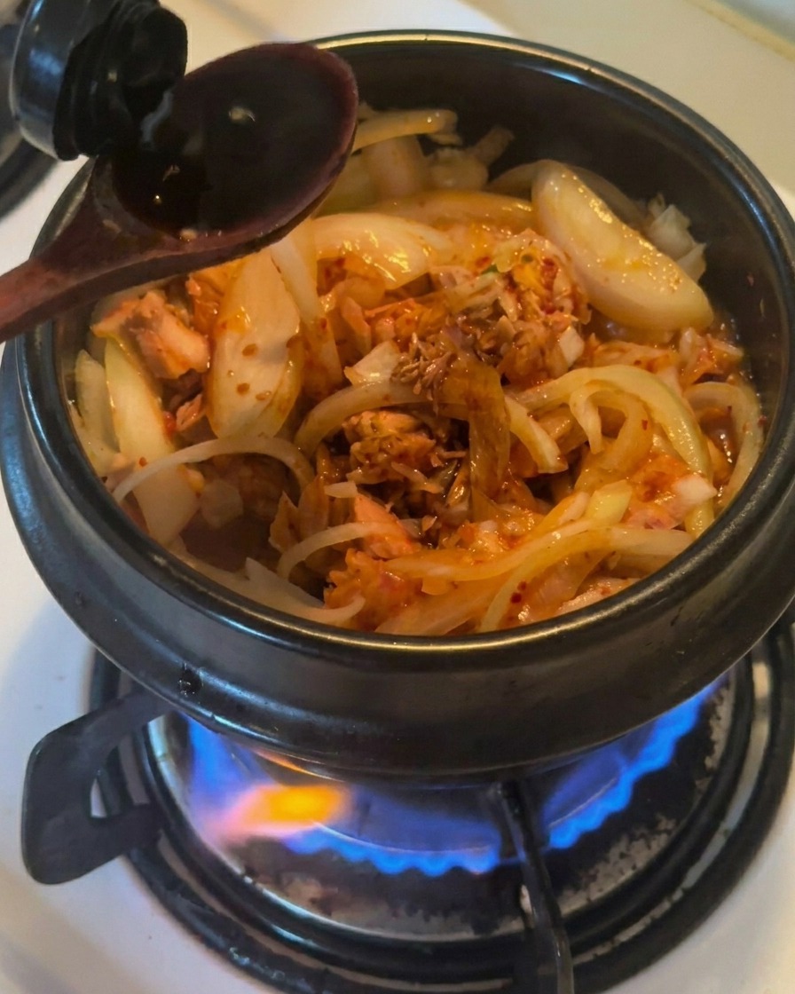 Une cuillère en bois versant de la sauce soja foncée dans une marmite en terre cuite noire contenant les oignons, le thon et le kimchi.