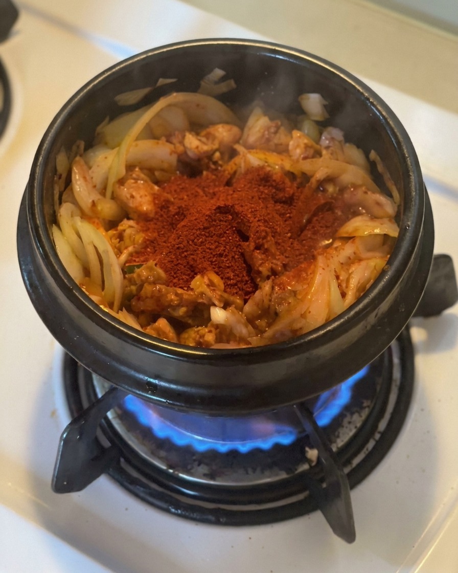 Une généreuse dose de gochugaru rouge vif posée sur le kimchi et les oignons sautés dans une marmite en pierre noire.