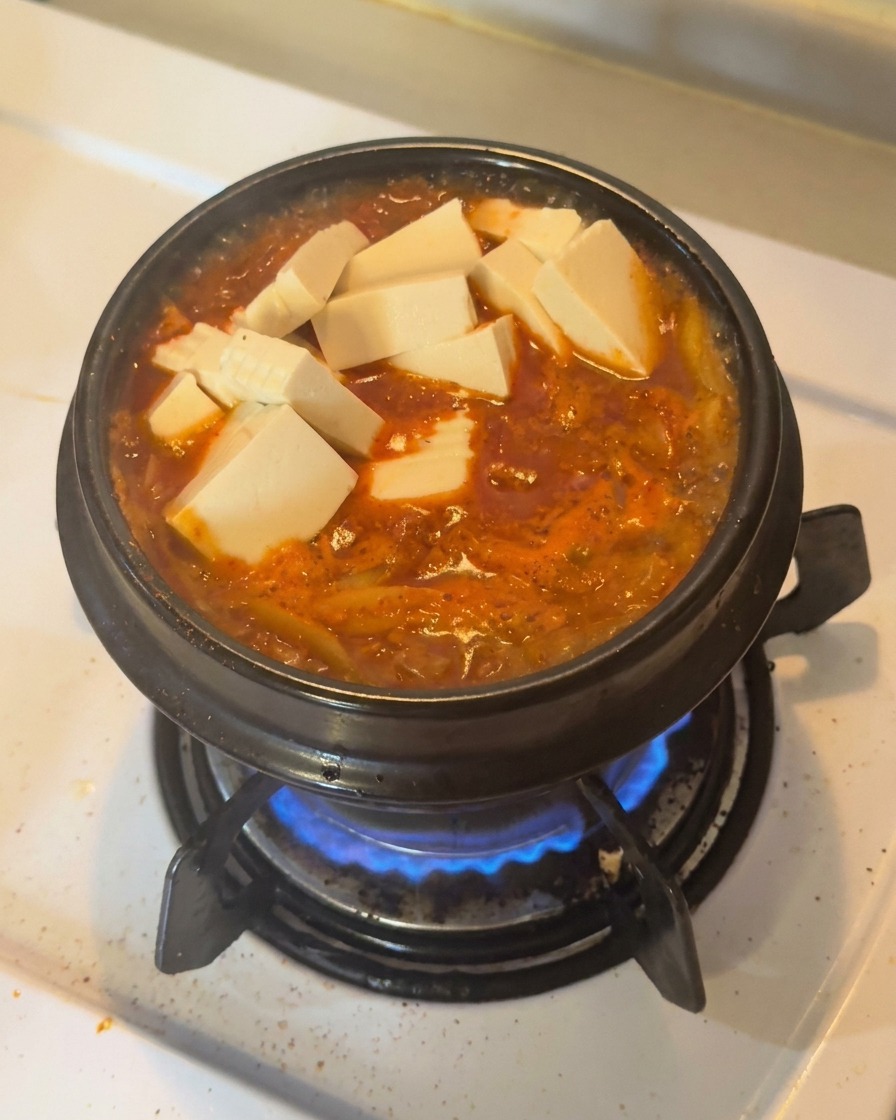 Cubes de tofu blanc frais et rondelles de piments verts disposés sur un ragoût de kimchi bouillonnant dans une marmite en terre.