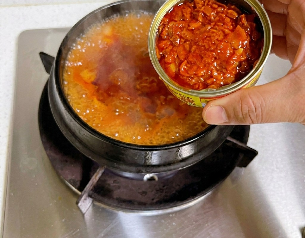Une main tenant une boîte de conserve ouverte et versant des morceaux de thon dans un bouillon de kimchi rouge frémissant.