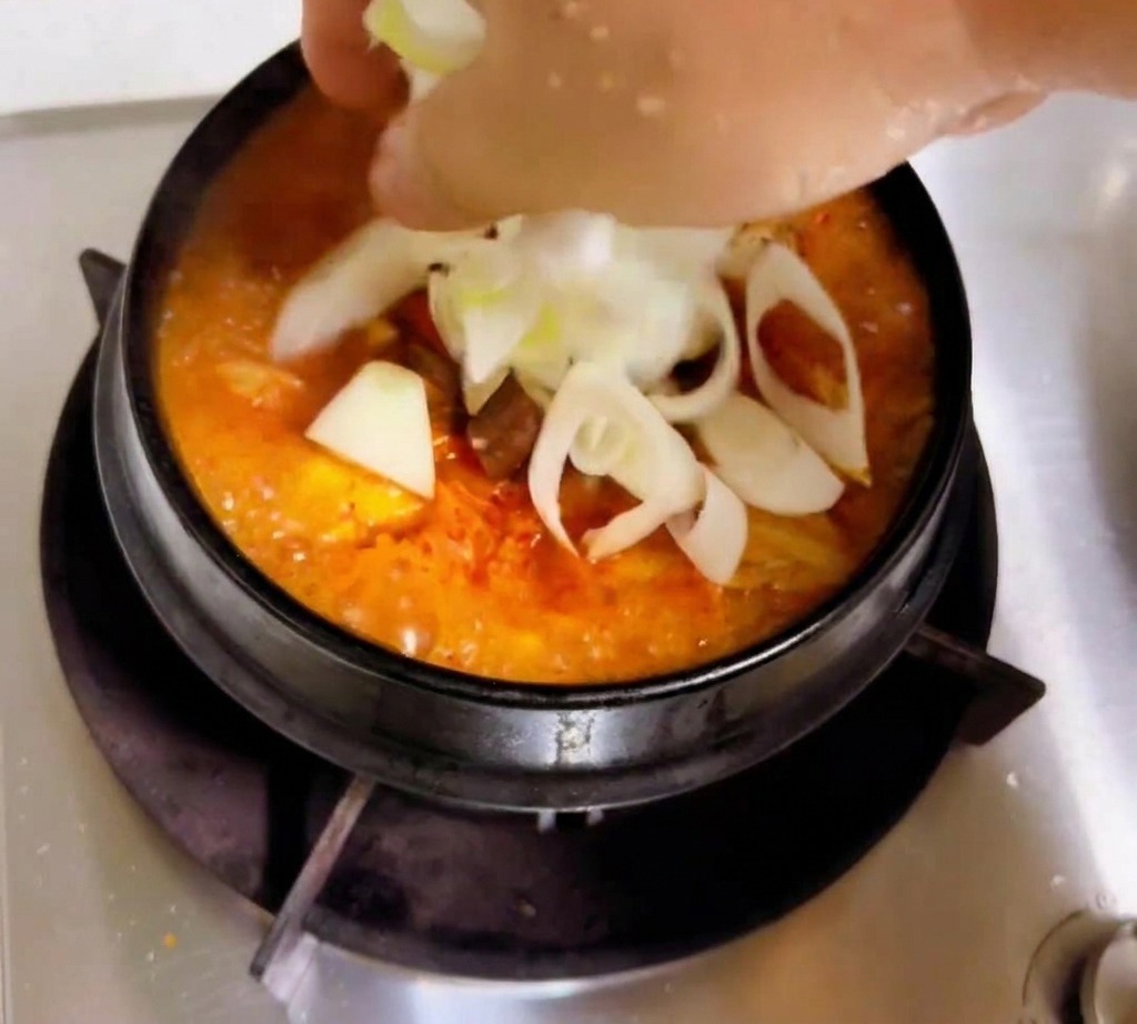 Une main ajoutant des rondelles doignons blancs et des oignons verts à un ragoût de kimchi orange-rouge qui mijote.