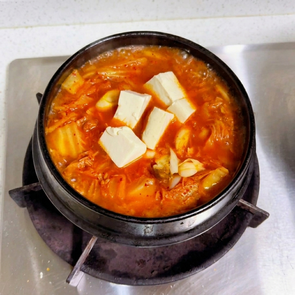 Cubes de tofu blanc frais reposant sur un bouillon de kimchi rouge bouillonnant dans un pot noir.