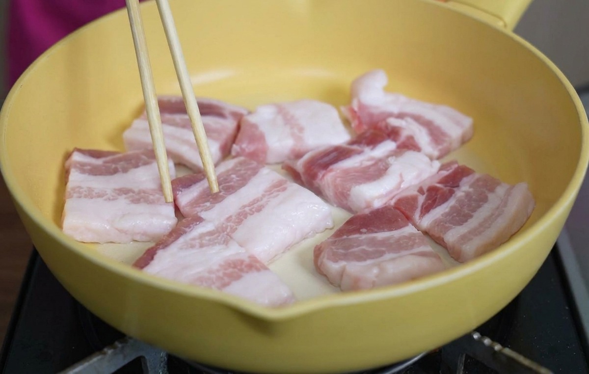 Tranches épaisses de poitrine de porc crue disposées dans une poêle en céramique jaune, ajustées avec des baguettes.
