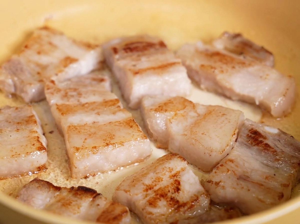 Tranches de poitrine de porc frites, bien dorées et croustillantes, grésillant dans leur graisse dans une poêle jaune.