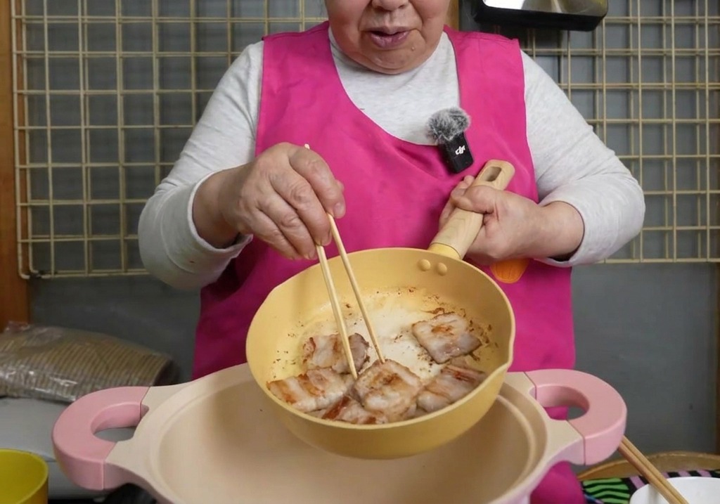 Tranches de poitrine de porc dorée transférées à laide de baguettes depuis une poêle jaune vers une grande marmite rose.