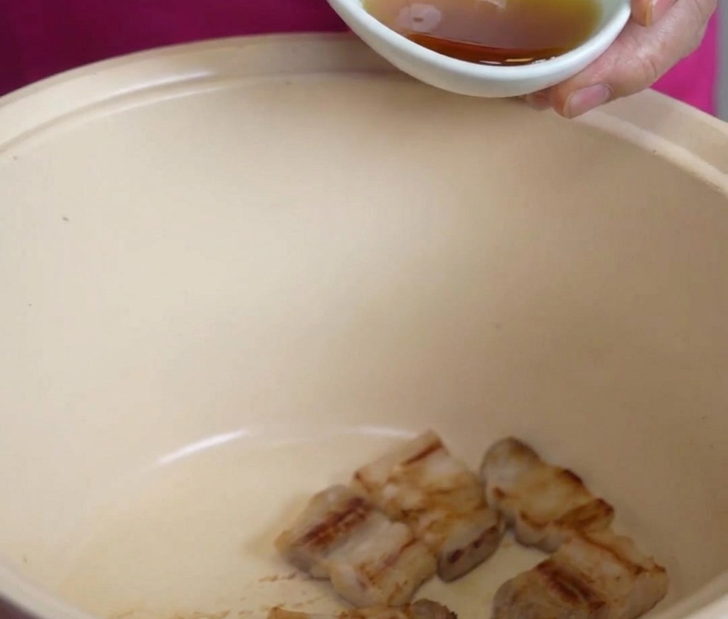 Un petit bol blanc versant de lhuile de sésame dorée sur des morceaux de poitrine de porc frite dans une grande marmite de couleur crème.