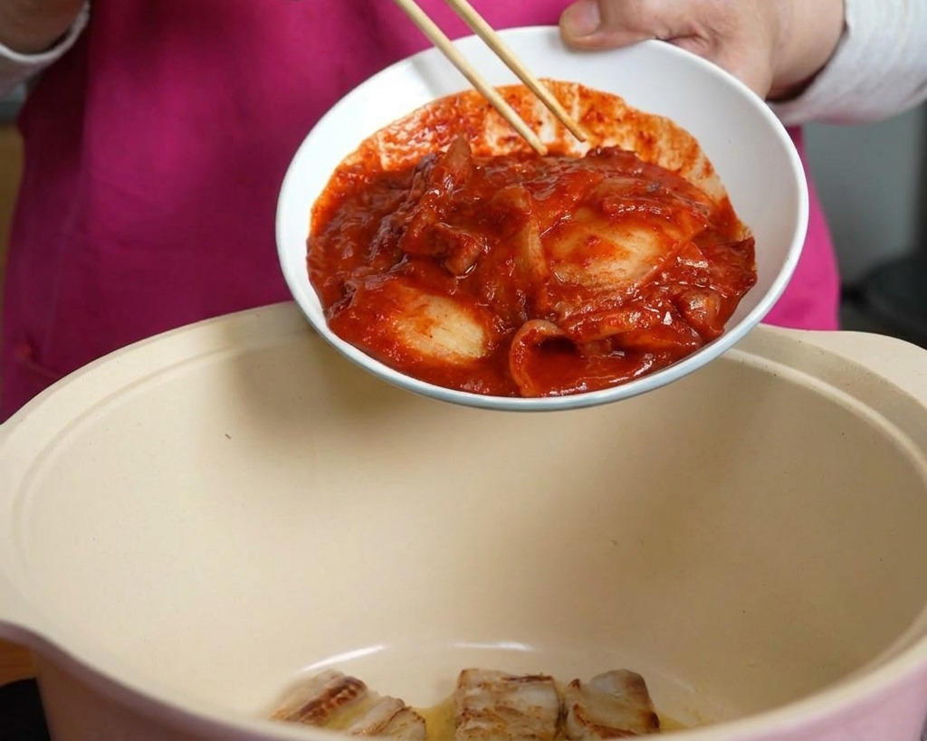 Une main utilisant des baguettes pour faire glisser un bol de kimchi rouge épicé dans une marmite contenant la poitrine de porc.