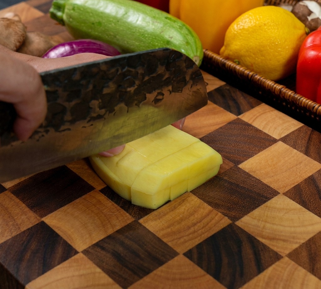 Un couteau de cuisine coupant une pomme de terre jaune épluchée en dés réguliers sur une planche en bois.