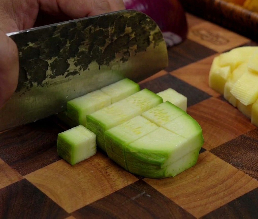 Un couperet coupant une courgette verte en petits morceaux rectangulaires sur une planche en bois quadrillée.