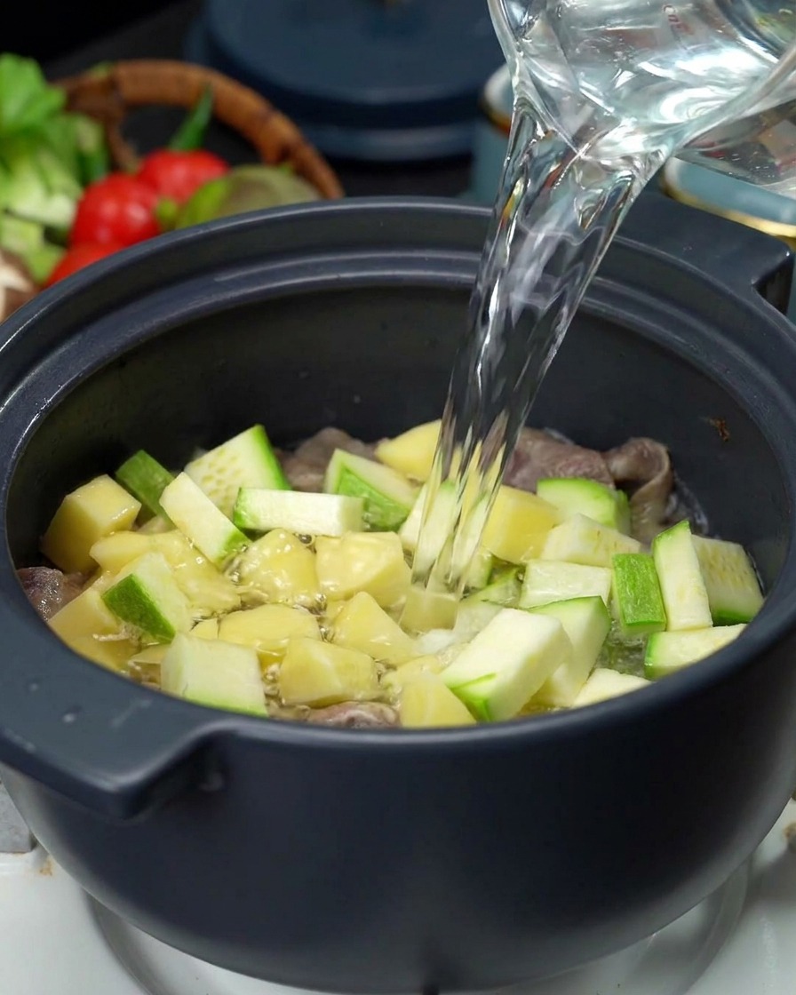 Un filet deau claire versé depuis une carafe en verre dans une marmite remplie de viande, de pommes de terre et de courgettes.