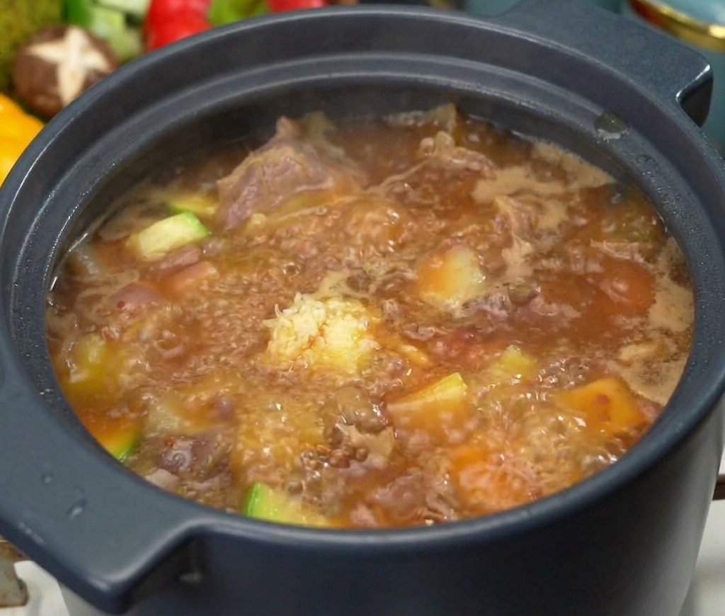 Une cuillère dail haché posée au-dessus dune marmite frémissante de soupe coréenne à la pâte de soja et au bœuf, avec des morceaux de légumes visibles.