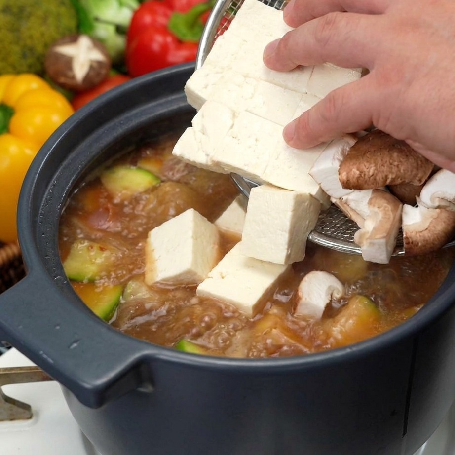 Une main utilisant une écumoire pour ajouter des dés de tofu blanc frais et des tranches de champignons shiitake bruns dans la soupe de soja frémissante.