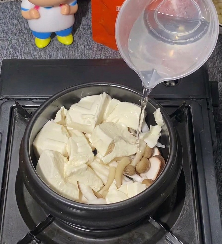 De leau claire versée depuis un verre doseur en plastique dans une marmite noire remplie de tofu et de champignons.