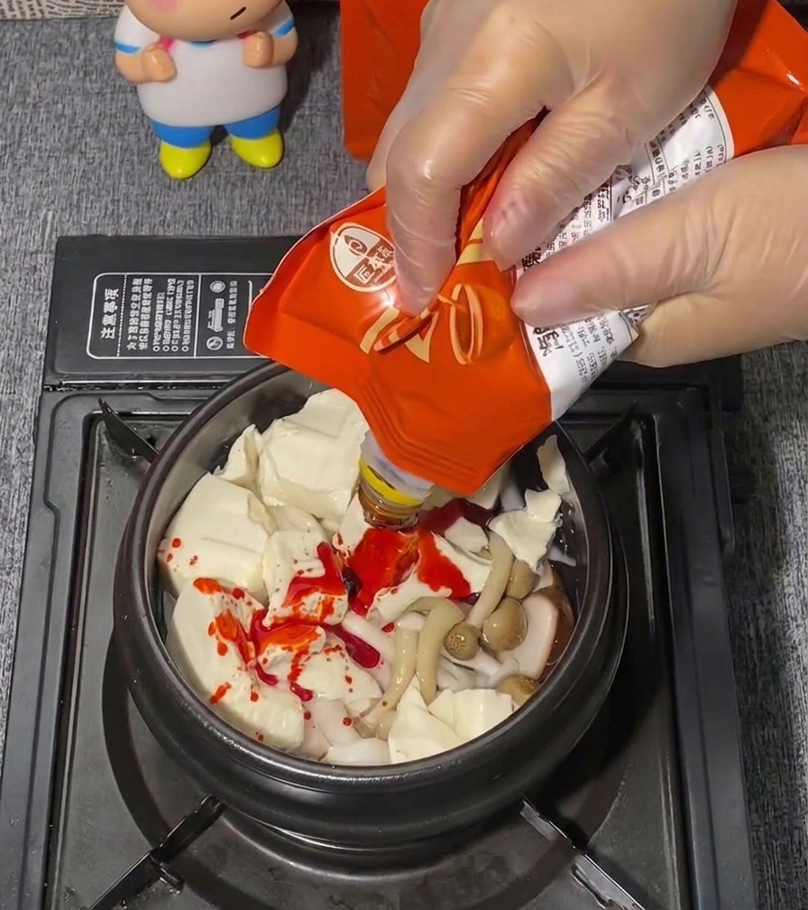 Une main portant un gant en plastique transparent pressant une pâte dassaisonnement rouge vif depuis un sachet sur un lit de tofu soyeux.