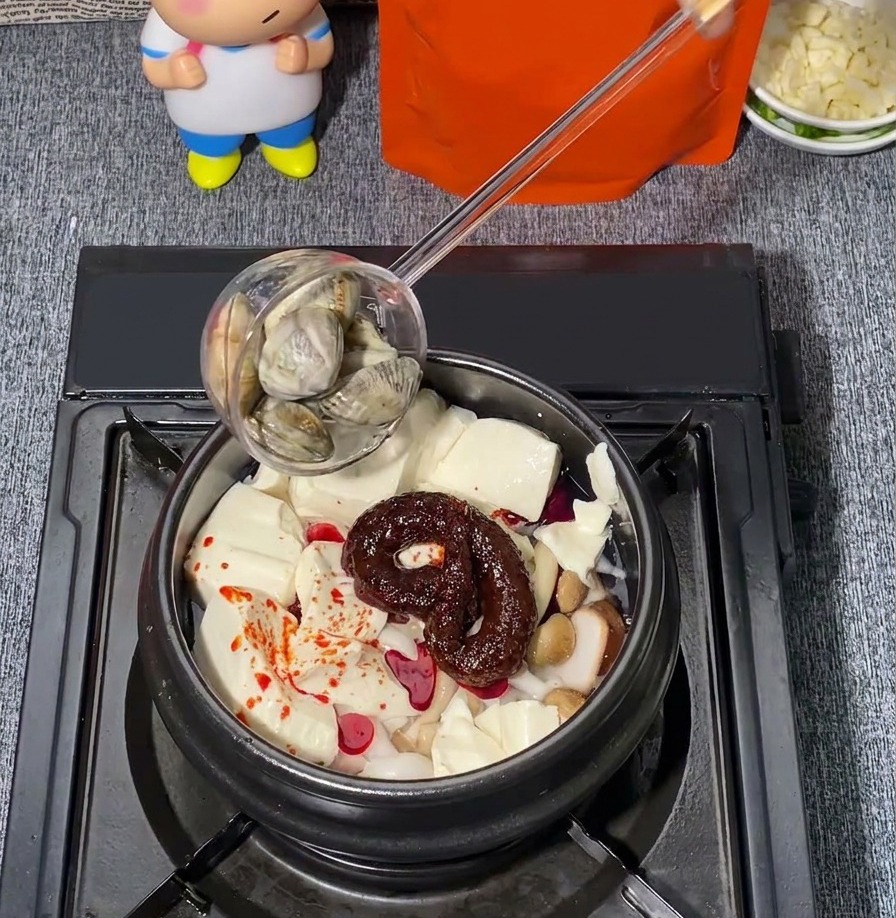 Des palourdes fraîches ajoutées à laide dune louche dans un pot en pierre contenant du tofu soyeux, des champignons et un assaisonnement au piment rouge.
