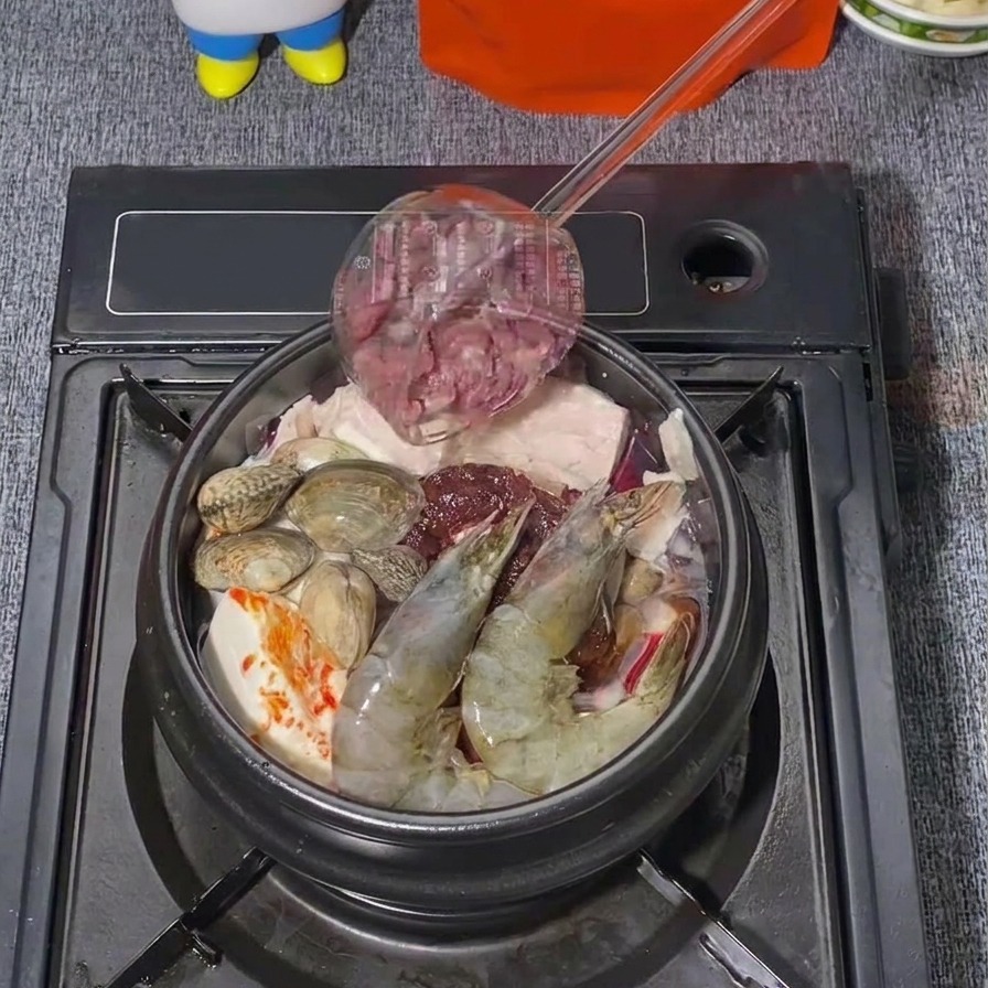 De fines tranches de bœuf cru ajoutées depuis une louche dans une marmite de ragoût coréen contenant déjà des crevettes et des palourdes.