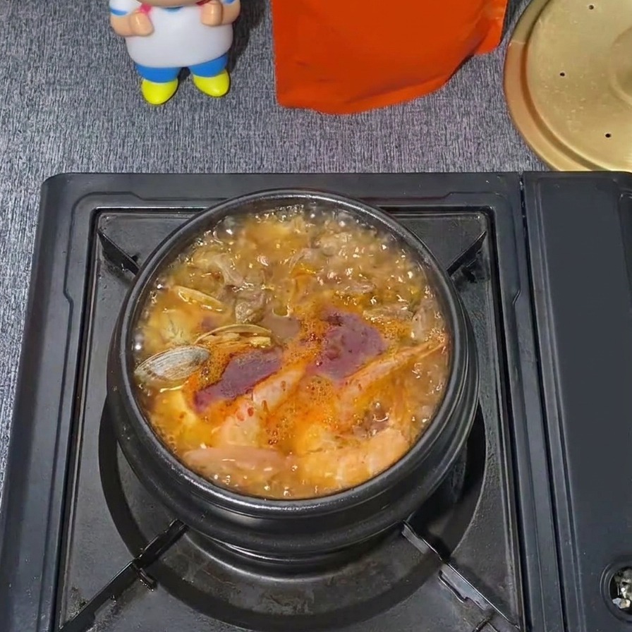 Une marmite frémissante de ragoût de tofu soyeux coréen aux fruits de mer avec un bouillon rouge vif, des palourdes visibles et de la vapeur qui séchappe.