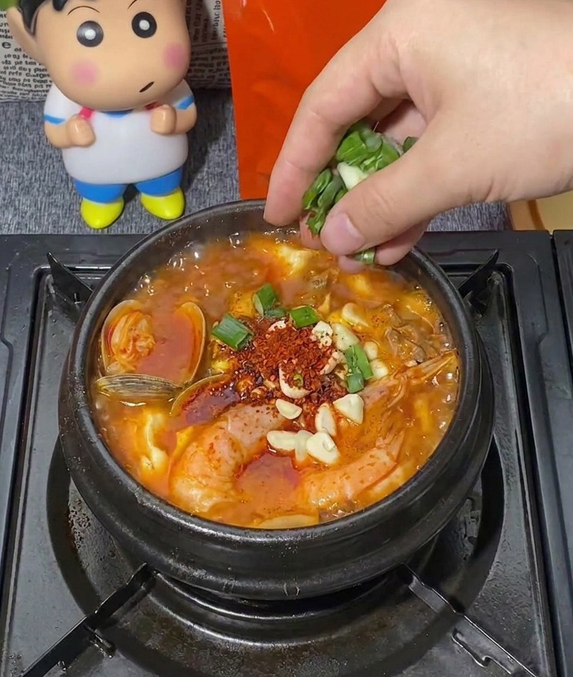 Une main parsemant une poignée de cébettes émincées dans un pot en pierre noir frémissant de ragoût coréen garni de crevettes, palourdes et ail haché.