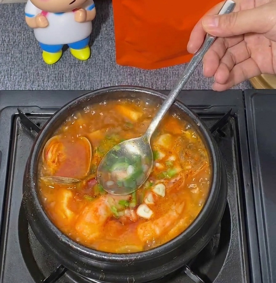 Une cuillère en métal mélangeant des oignons verts émincés dans un pot frémissant de ragoût de tofu soyeux rouge aux fruits de mer.