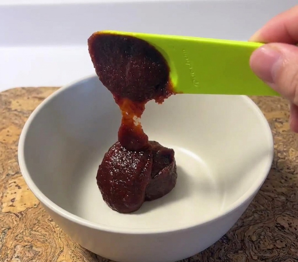 Une cuillérée de pâte de piment coréenne rouge foncé et épaisse déposée dans un bol en céramique blanc avec une cuillère verte