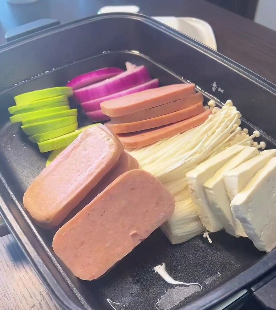 Une marmite noire garnie de sections soigneusement alignées doignon rouge, de courgettes, de jambon en conserve, de champignons enoki et de blocs de tofu.