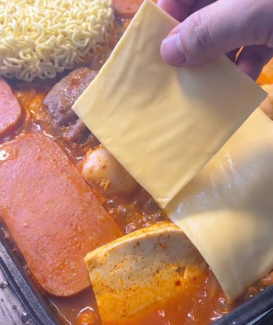 Une main déposant une tranche carrée de fromage jaune sur des nouilles instantanées au cœur dun ragoût rouge bouillonnant.