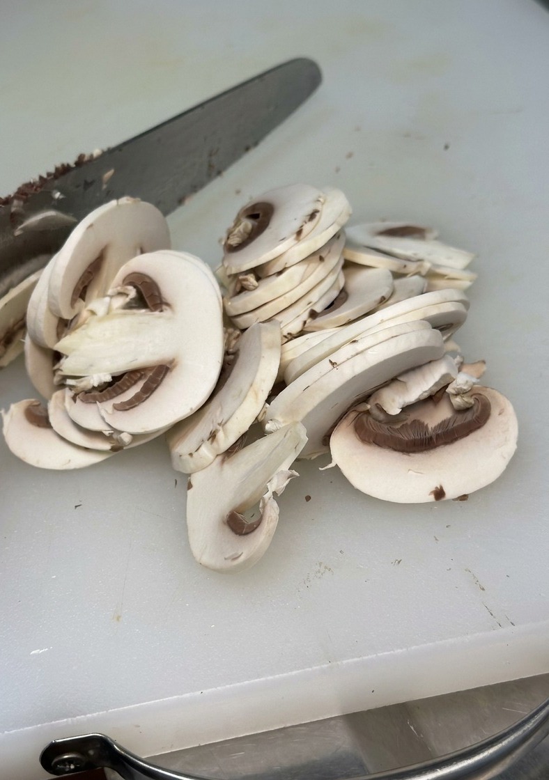 Gros plan dun champignon blanc émincé en fines tranches régulières avec un couteau métallique sur une planche à découper blanche.