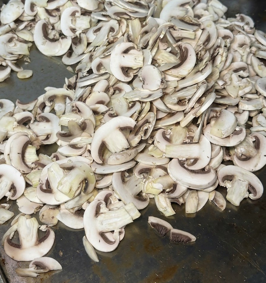 Fines tranches de champignons étalées sur une plancha métallique chaude, dégageant de la vapeur au début de la cuisson.