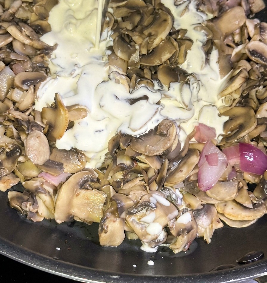 Un filet de crème fraîche liquide coulant dune brique bleue dans une poêle remplie de champignons sautés et doignons rouges en dés.