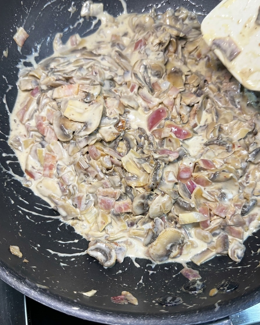 Une spatule blanche remuant une sauce riche et crémeuse remplie de tranches de champignons, doignons rouges en dés et de morceaux de lardons dans une poêle noire.