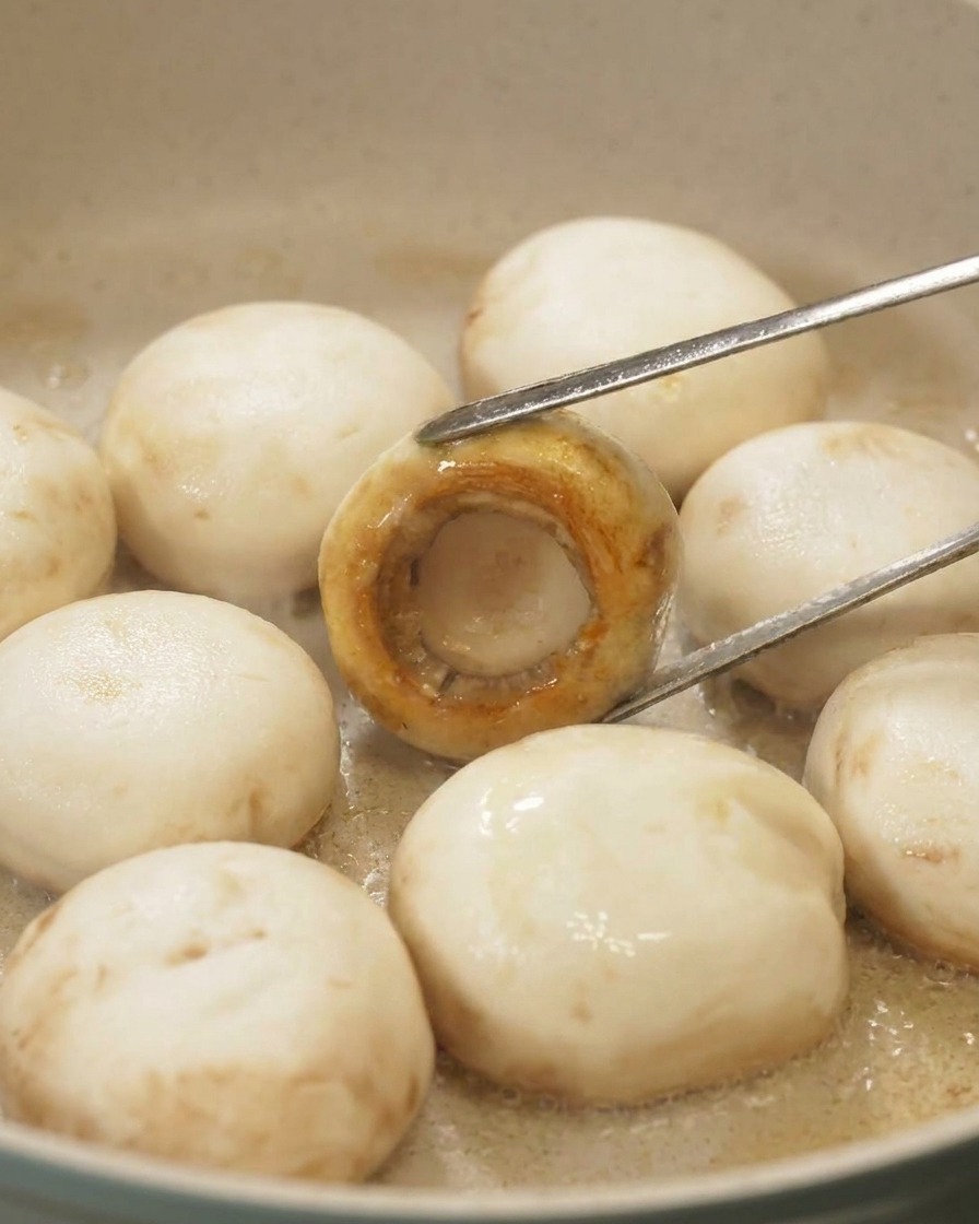 Une pince retournant un champignon dans une poêle pour révéler une surface parfaitement caramélisée et dorée sur le chapeau.