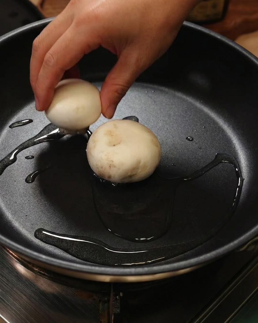 Une main déposant un champignon farci de pâte de crevettes, face farce vers le bas, dans une poêle contenant de lhuile.