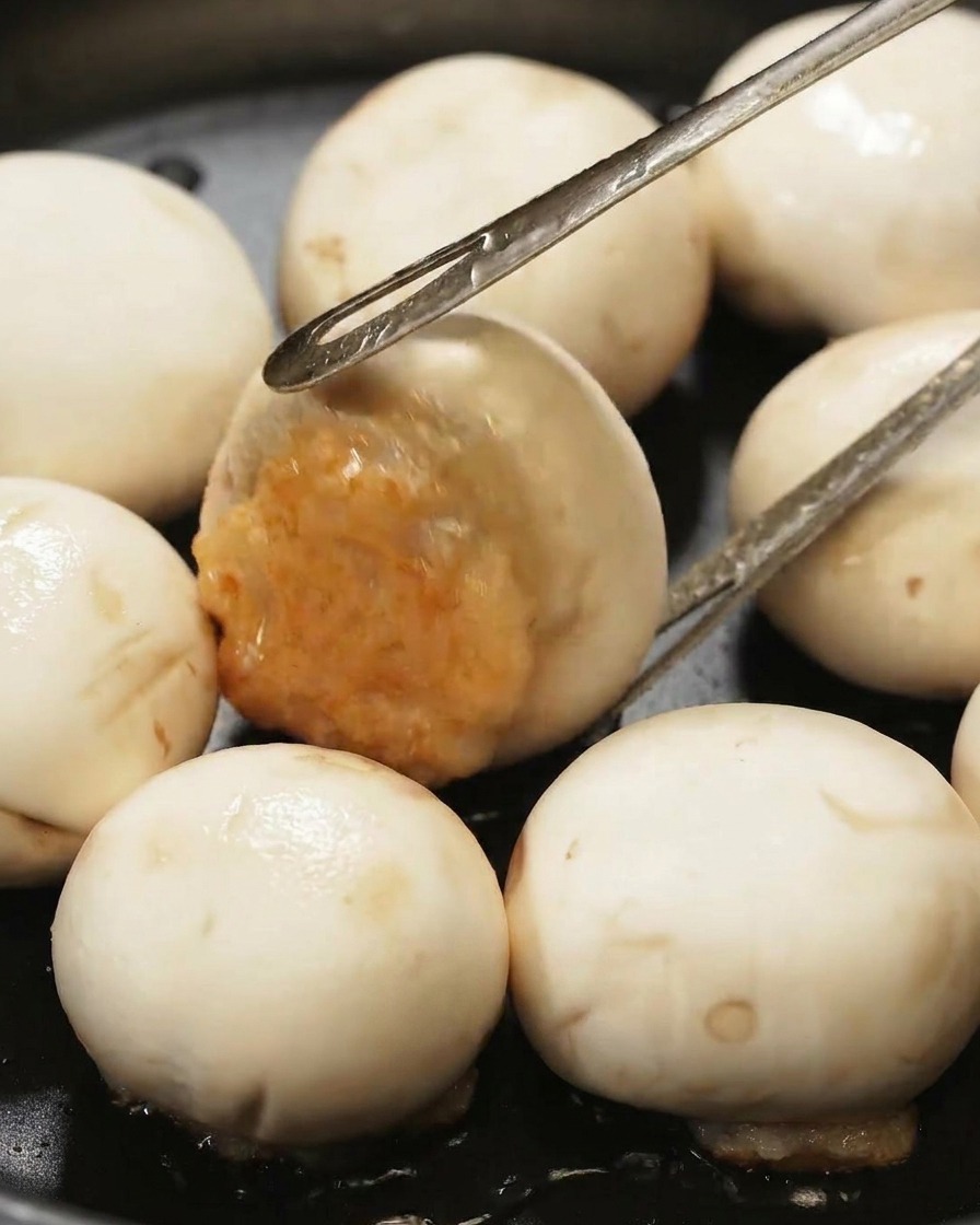 Une pince métallique retournant un champignon farci dans une poêle noire pour révéler la farce de crevettes bien dorée.