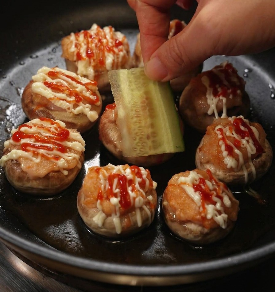 Une main déposant une fine rondelle de concombre frais sur un mini-burger de champignon farci nappé de mayonnaise et de ketchup.