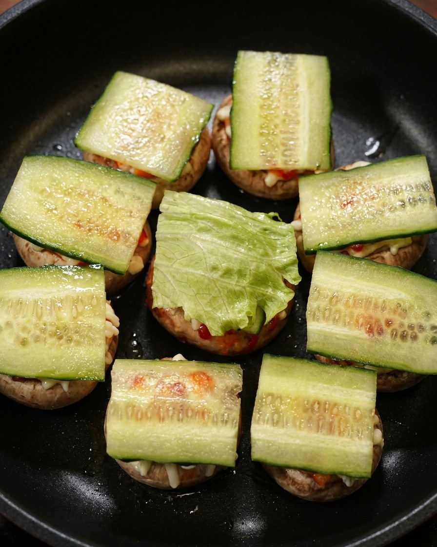 Des tranches de concombre et des feuilles de laitue verte superposées sur des mini-burgers de champignons dans une poêle noire.