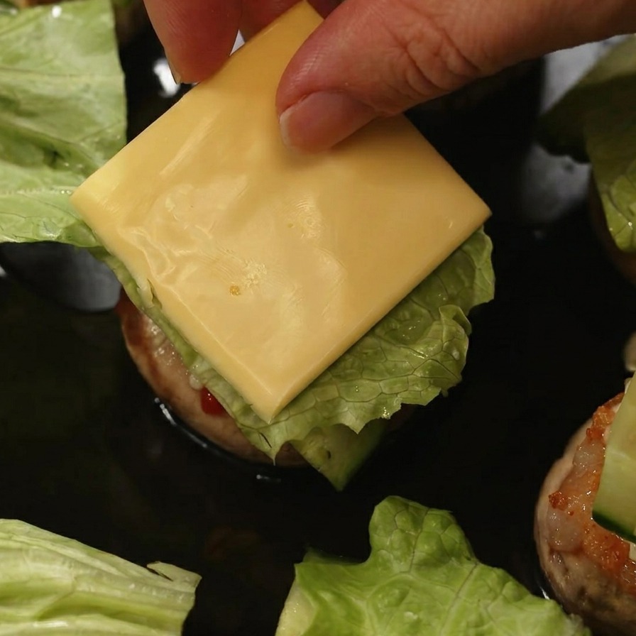 Une main déposant un carré de fromage jaune sur de la laitue et du concombre à lintérieur dun burger de champignon.