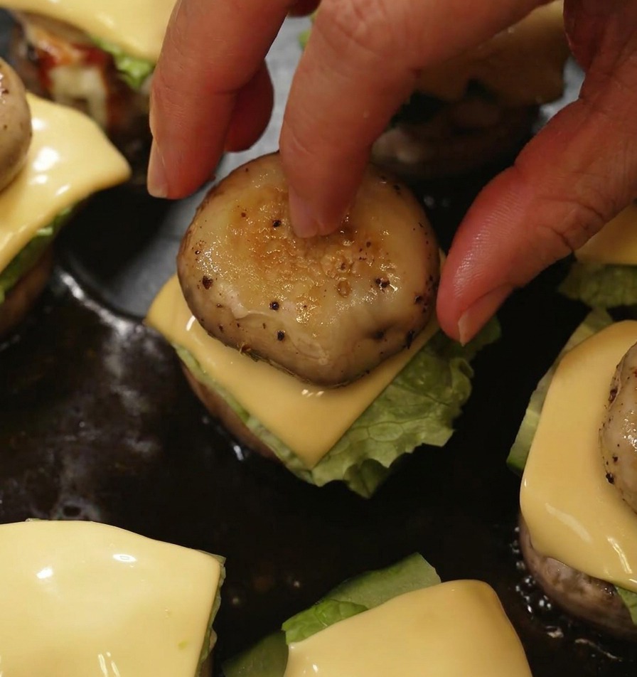 Une main posant un chapeau de champignon grillé sur un mini-burger pour terminer lassemblage dans la poêle.